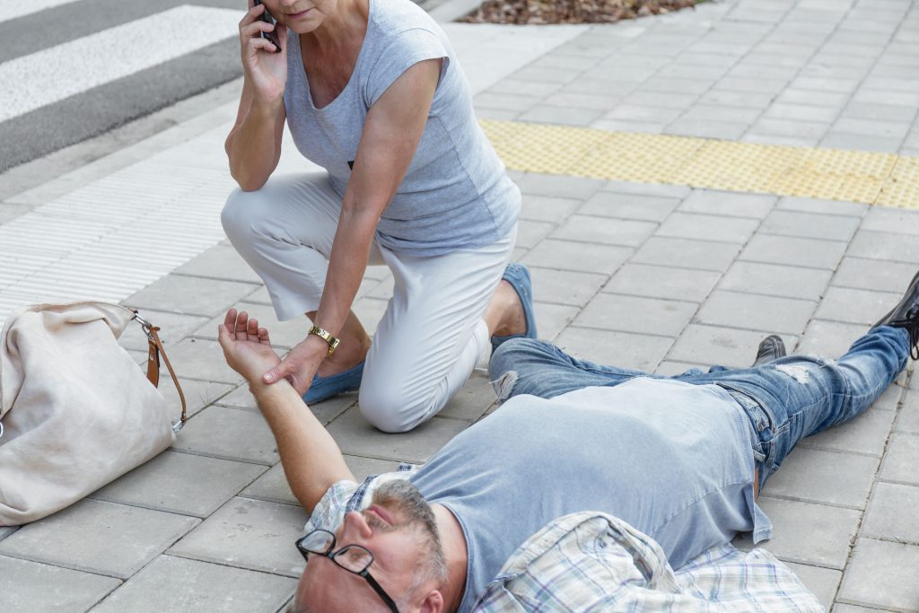 Senior woman calling emergency hotline while checking a man's vital functions on street.
